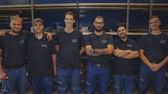 Die Gruppe von sechs Handwerkern posiert freundlich in blauen Uniformen, umgeben von Rohren und Werkzeugen in einem Arbeitsumfeld.