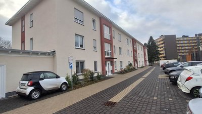 Eine moderne Wohnanlage mit mehreren Etagen, umgeben von einem gepflasterten Parkplatz und verschiedenen Autos.