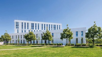 Ein modernes, weißes Gebäude mit großen Fenstern, umgeben von grünen Bäumen und einem klaren blauen Himmel.