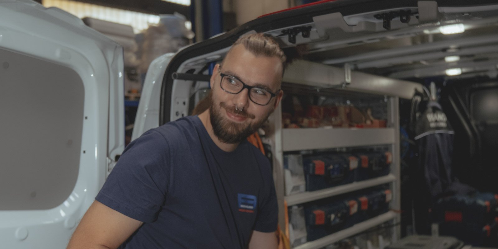 Ein junger Mann mit Bart und Brille lächelt, während er in einem Lieferwagen steht, umgeben von Werkzeugen und Ausrüstung.