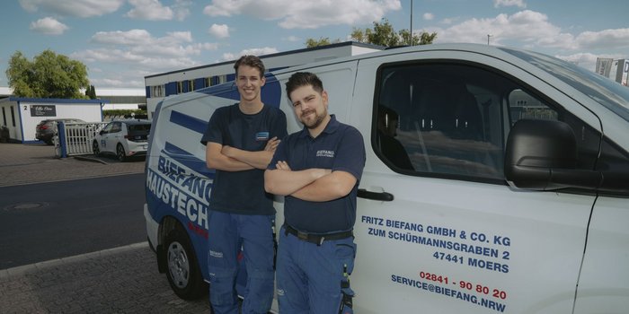 Zwei Männer in Arbeitskleidung stehen lächelnd vor einem Firmenfahrzeug, umgeben von einem klaren Himmel und modernen Gebäuden.