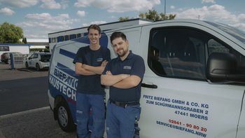 Zwei Männer in Arbeitskleidung stehen lächelnd vor einem Firmenfahrzeug, umgeben von einem klaren Himmel und modernen Gebäuden.