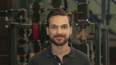 Ein Mann mit einem gepflegten Bart und einem freundlichen Lächeln steht vor einer industriellen Kulisse mit Maschinen.