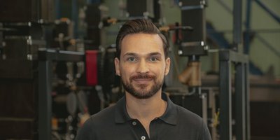 Ein Mann mit einem gepflegten Bart und einem freundlichen Lächeln steht vor einer industriellen Kulisse mit Maschinen.