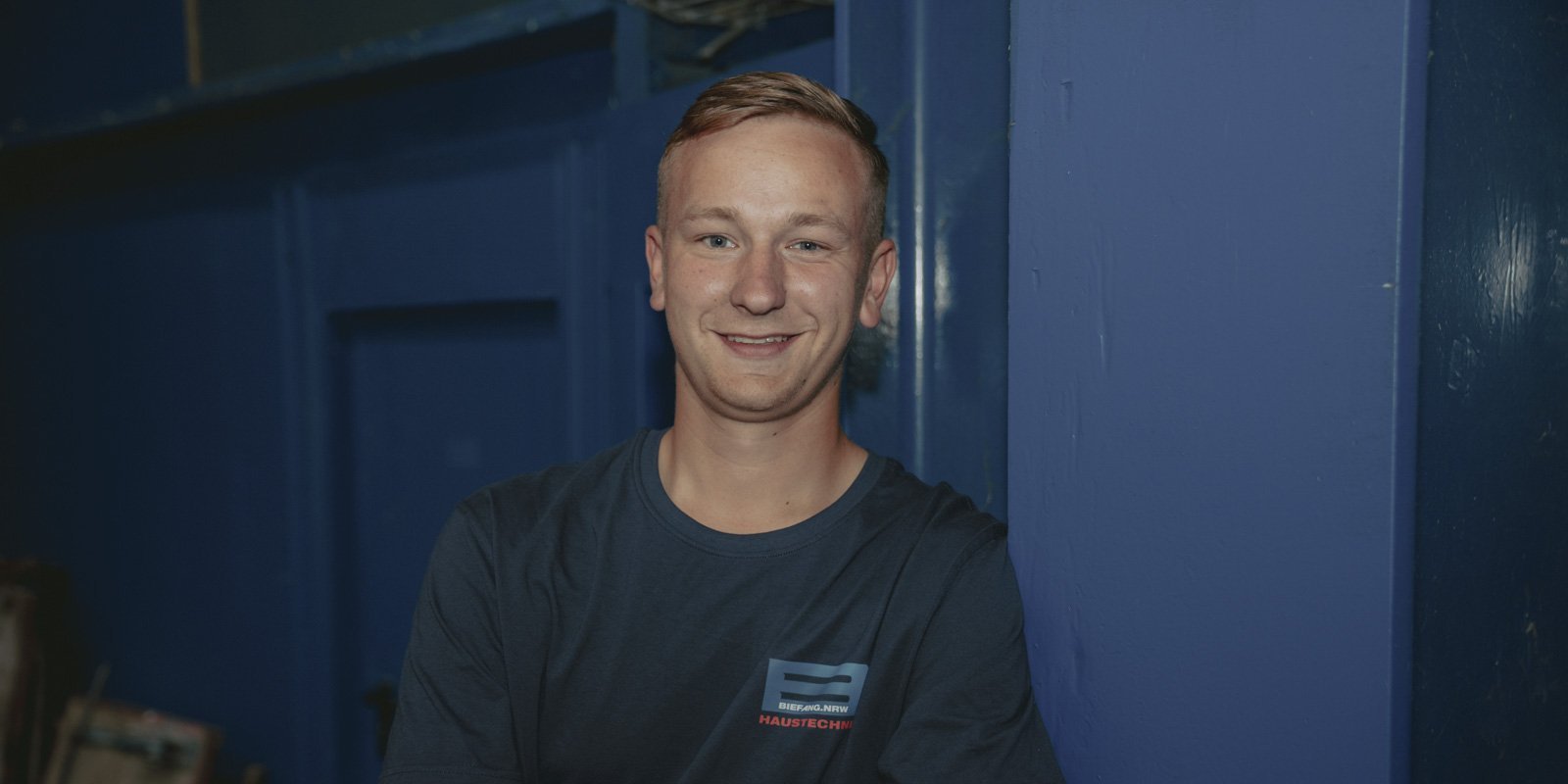 Ein junger Mann mit kurzem Haar lächelt entspannt, während er an einer blauen Wand lehnt, in einem gut beleuchteten Raum.
