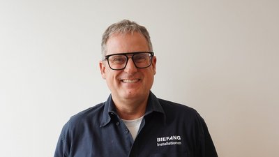Ein lächelnder Mann mit Brille trägt ein dunkles Hemd mit dem Aufdruck 'BIEFANG Installationen' vor einer hellen Wand.