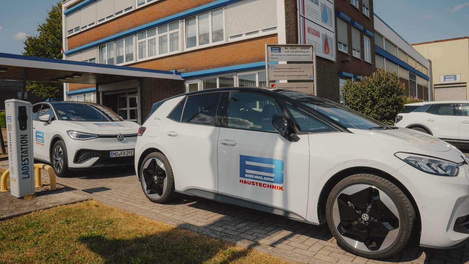 Zwei Elektroautos parken an einer Ladestation vor einem modernen Gebäude mit großen Fenstern und einem blauen Akzent.