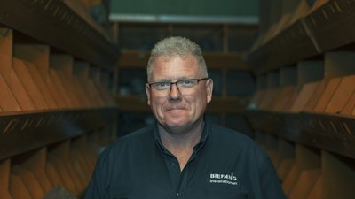 Ein Mann mit Brille und einem freundlichen Lächeln steht in einem Raum mit Regalen im Hintergrund, die orangefarbene Fächer zeigen.