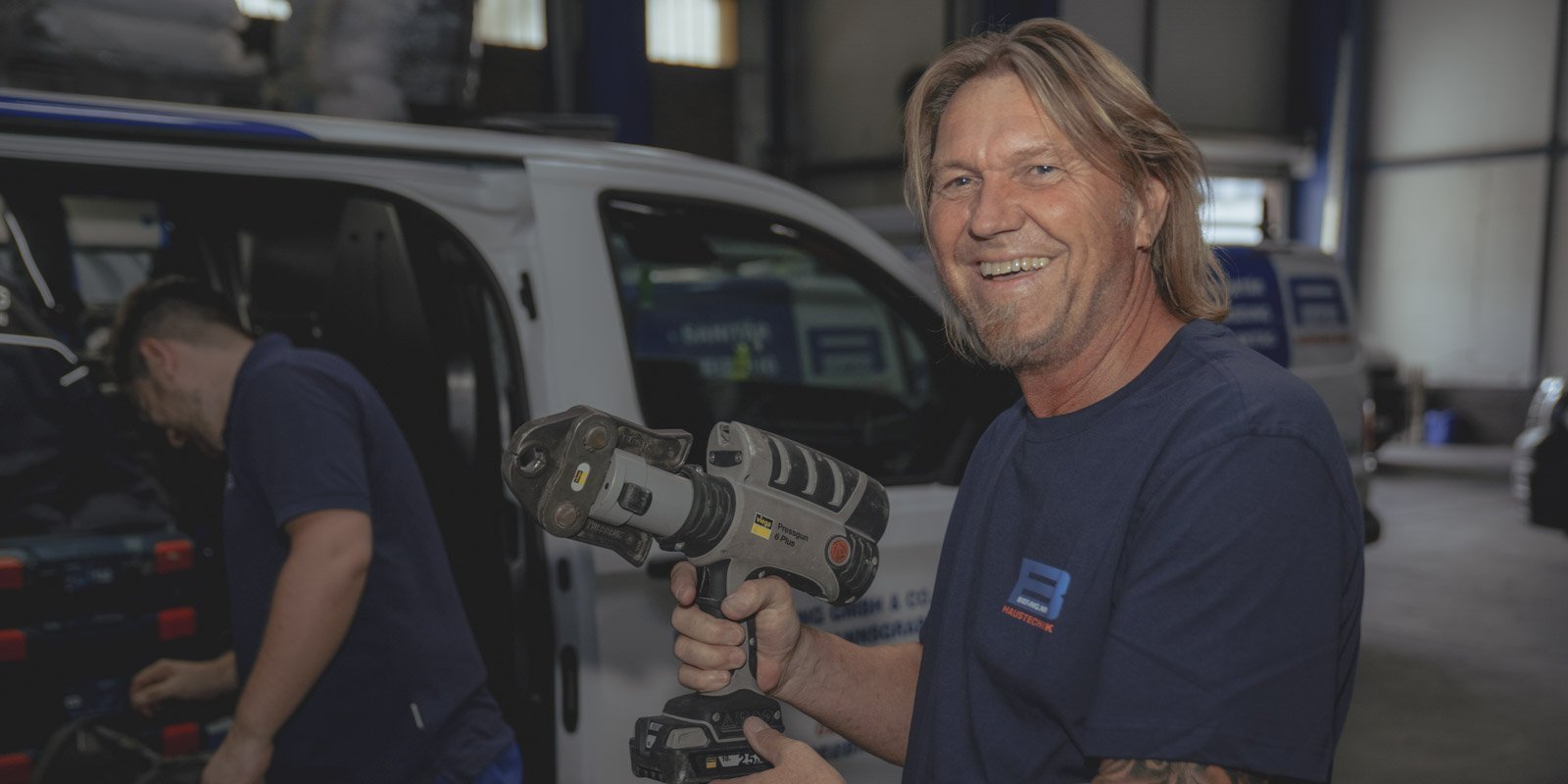 Ein lächelnder Mann hält ein Elektrowerkzeug in einer Werkstatt, während ein Kollege im Hintergrund arbeitet.