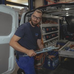 Ein Mechaniker mit einer Zange steht in einem Lieferwagen, umgeben von Werkzeugen und Wasserflaschen.