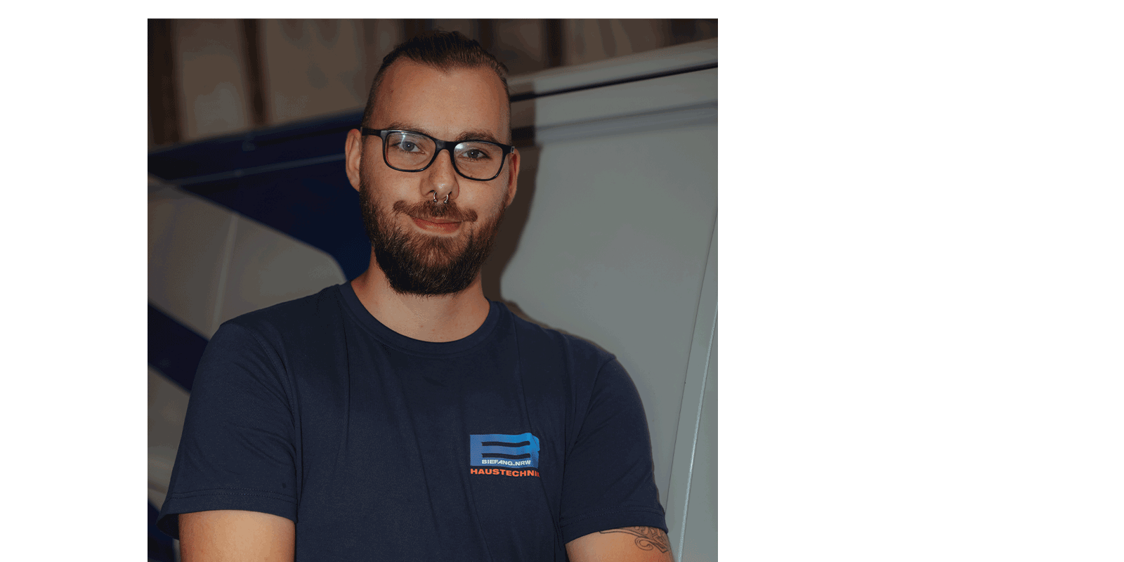 Ein junger Mann mit Bart und Brille lächelt selbstbewusst, während er in einem technischen Umfeld steht.