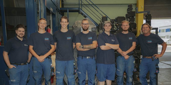 Eine Gruppe von sieben Männern in blauen Arbeitskleidung steht lächelnd in einer Werkstatt, umgeben von Maschinen und Werkzeugen.