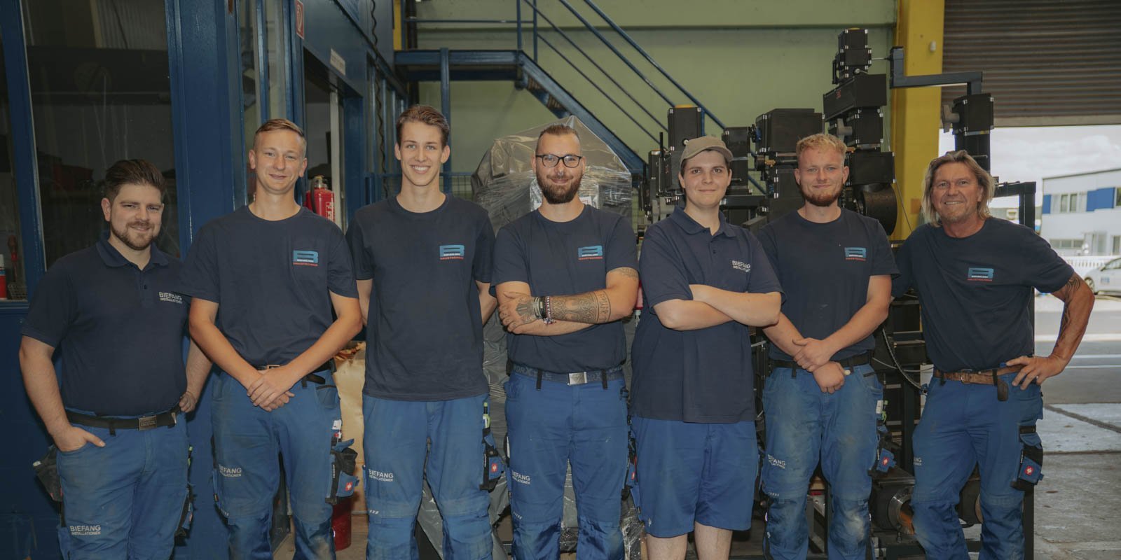 Eine Gruppe von sieben Männern in blauen Arbeitskleidung steht lächelnd in einer Werkstatt, umgeben von Maschinen und Werkzeugen.