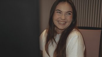 Eine junge Frau mit langen Haaren lächelt fröhlich, während sie an einem Schreibtisch sitzt, umgeben von einem modernen Raum.