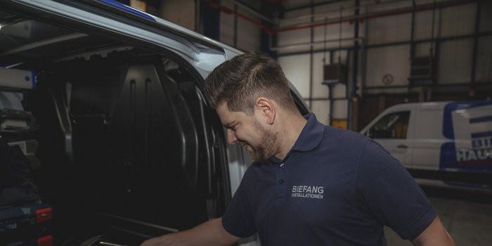 Ein junger Mann mit einem freundlichen Lächeln schaut in den Kofferraum eines Fahrzeugs, um Ausrüstung zu überprüfen.