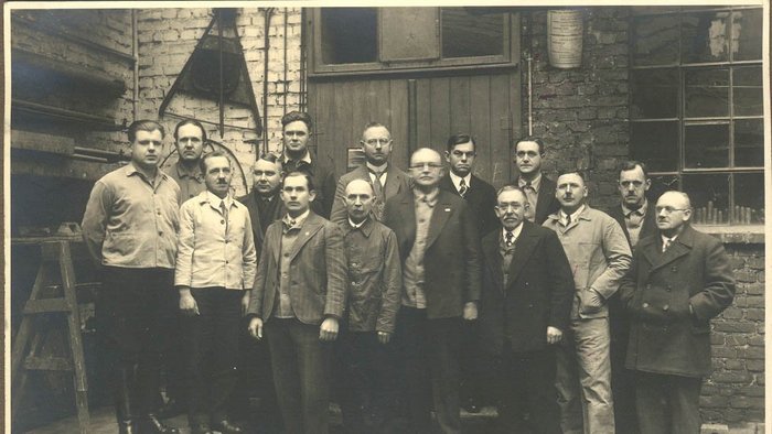 Eine Gruppe von Männern steht in einer Werkstatt, umgeben von Ziegelwänden und Werkzeugen, alle in formeller Kleidung.