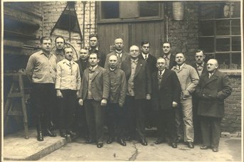Eine Gruppe von Männern steht in einer Werkstatt, umgeben von Ziegelwänden und Werkzeugen, alle in formeller Kleidung.