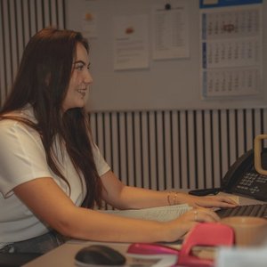 Eine junge Frau mit langen Haaren lächelt, während sie an einem Schreibtisch mit einem Telefon und Notizen arbeitet.
