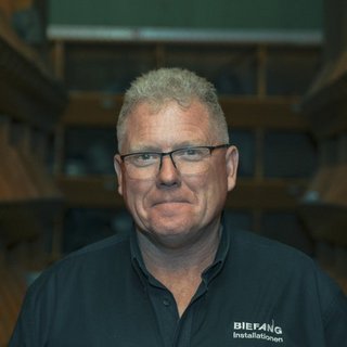Ein Mann mit Brille und kurzem, grauem Haar lächelt freundlich vor einem Holzregal im Hintergrund.