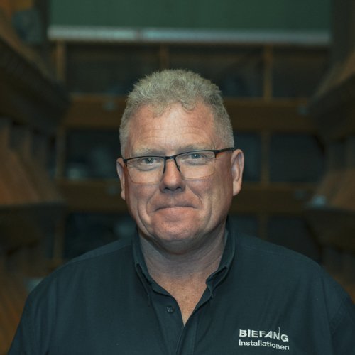 Ein Mann mit Brille und kurzem, grauem Haar lächelt freundlich vor einem Holzregal im Hintergrund.